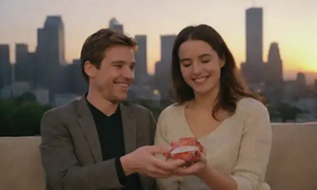 2 personas abrazando, a mano, sonriendo unos a otros, con un paisaje urbano o fondo de puesta de sol; o una pareja sentada en un sofá, riéndose juntos, con un enfoque sutil en una persona que sostiene un pequeño regalo o sorpresa para el otro, transmitiendo confianza y prioridad