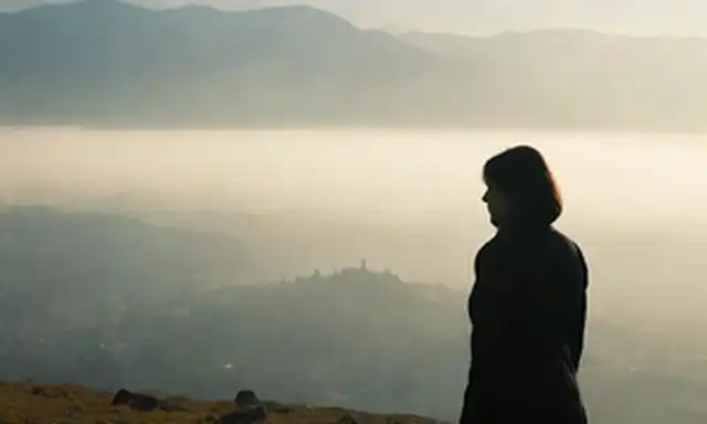 Una figura de pie sola en la cima de una montaña, mirando hacia un vasto paisaje; una fuente de luz cálida que arroja largas sombras; una bruma sutil o niebla envolviendo la escena; un sentido de aislamiento y contemplación; un paisaje urbano distante o edificios en el fondo, borrosos o inmundos; un fuerte contraste entre la luz brillante y las sombras oscuras