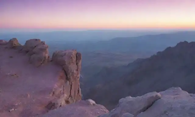 Un paisaje borroso y abstracto con nubes brillantes y una línea de horizonte débil, una figura solitaria de pie en un afloramiento rocoso mirando hacia atrás al espectador, un sutil gradiente de colores que cambia de azul a púrpura, enfoque suave en montañas distantes, indicio de una marca de interrogación en la distancia