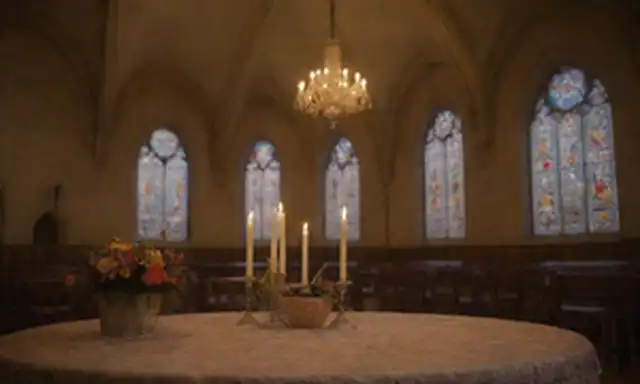 Habitación doble, suavemente enfocada en un mantel con patrones de encaje intrincados, rodeado de flores, cestas de Pascua, y una majestuosa fachada de la iglesia, luz caliente emanando de vidrieras, sombras echadas en las paredes