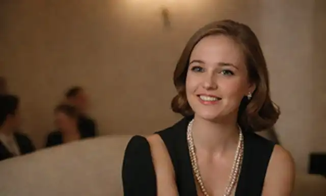 Mujer a principios de los años veinte con una sonrisa profesional, sentada elegantemente con las manos en la vuelta, con traje de negocios y perlas, fondo con sutil patrón floral, iluminación cálida