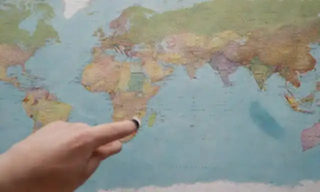 Un mapa con marcadores coloridos, una persona señalando diferentes lugares, banderas ondeando en el fondo, manos sujetando globos y brújulas, símbolos para idiomas y culturas, y un sutil gradiente sugiriendo movimiento y exploración
